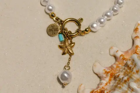 Un delicado llavero de oro con charms de estrella de mar y perla, sobre arena y junto a una concha marina.