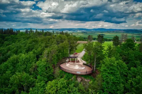 Widok na taras widokowy w otoczeniu lasu, z panoramą pól i gór w tle. Jakby to było jezioro wśród gór stołowych.