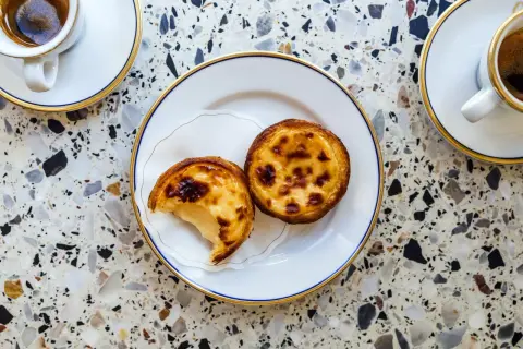 Dwa pyszne pastel de nata na talerzyku, obok filiżanki kawy. Idealny deser na każdą porę dnia.