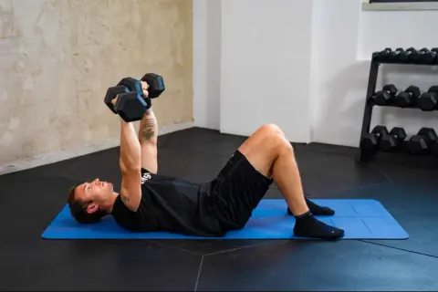 Uomo esegue esercizi pettorali con manubri su tappetino blu. Allenamento in palestra con rack di pesi sullo sfondo.