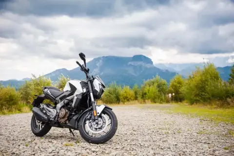 Biało-czarny motocykl Bajaj 400 na kamienistym podjeździe, w tle góry i chmury.