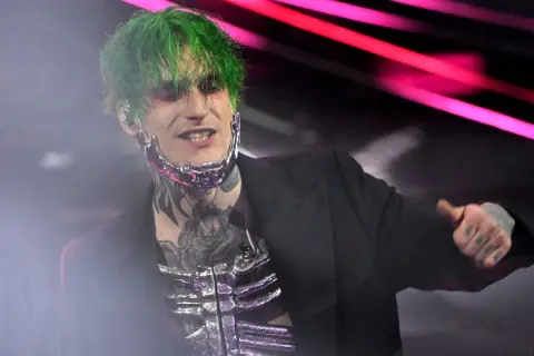 Theo la sad, con capelli verdi e trucco drammatico, indossa un abito nero e una maschera metallica.