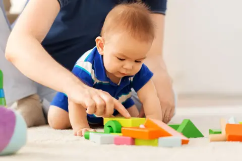 Dziecko w niebieskiej koszulce bawi się kolorowymi klockami na dywanie, a obok widać ręce dorosłego.