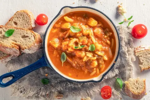 Herzhaftes Minestrone Rezept in einer blauen Pfanne, garniert mit Basilikum. Dazu gibt es knuspriges Brot und frische Tomaten.