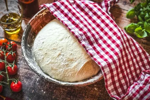 Ein Laib bester italienischer pizzateig ruht in einer Schüssel, bedeckt mit einem rot-weiß karierten Tuch. Daneben liegen frische Tomaten und Basilikum.