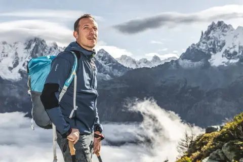 Randonneur souriant, sac à dos bleu et bâtons de marche, prêt à affronter le poids de la montagne. Paysage alpin spectaculaire.
