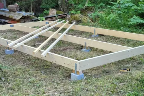 Grundläggande stomme för att bygga hus på plintar. Träbjälkar vilar på betongblock i en skogsmiljö.