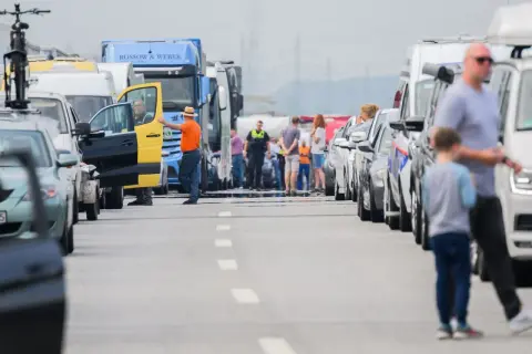 Stau auf der Autobahn. Ein Mann in orangefarbener Jacke und Hut steht neben einem gelb-schwarzen Transporter, dessen Tür offen ist. Viele Autos und LKW stehen still, als ob sie trotz Fahrverbot unterwegs wären.