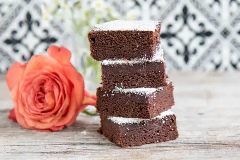 Schokoladige Brownies mit Datteln, bestäubt mit Puderzucker, neben einer zarten orangefarbenen Rose auf Holztisch.