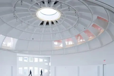 The expansive, white interior of the Faena Art Project Room features a striking domed ceiling with a central skylight. Two figures walk through the minimalist space.