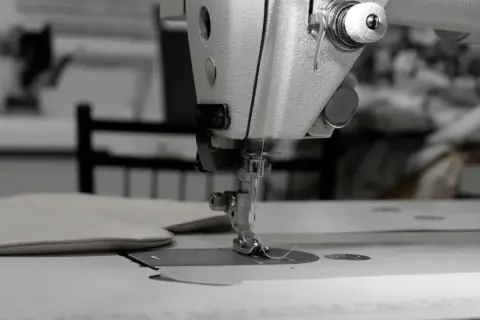 Zbliżenie na maszynę do szycia, która pracuje nad jasną, pętelkową tkaniną. W tle widać inne maszyny i materiały.