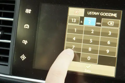 Palec dotyka ekranu podczas ustawiania godziny w Opel Astra H. Wyświetlacz pokazuje klawiaturę numeryczną do wprowadzania czasu.