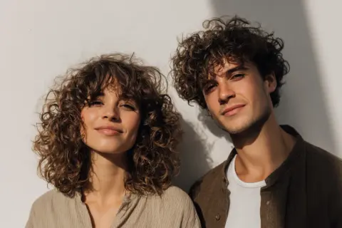 Ein junges Paar mit vollen Locken vor einer weißen Wand. Die curly girl lächelt sanft.