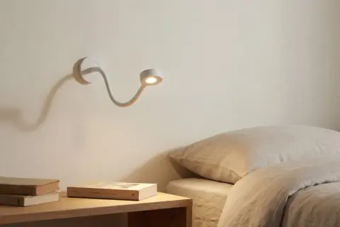Biała, elastyczna lampa nocna oświetla stos książek na stoliku obok łóżka.