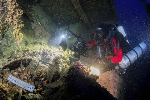 Nurkowie badają wrak statku, być może **Britannic**, z kamerą i oświetleniem. Widoczne są szczątki, muszle i ciemne głębiny.