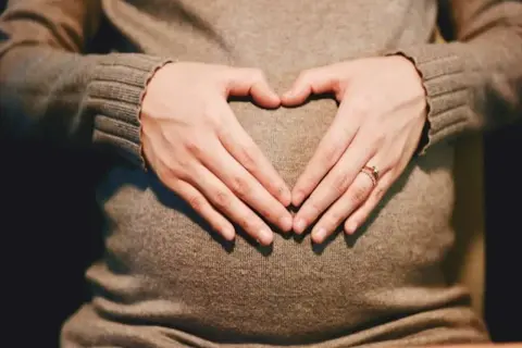 Dłonie tworzą serce na twardym brzuchu w ciąży. Widać obrączkę na palcu.