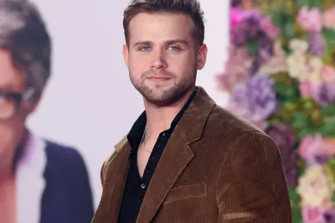 Leo Woodall, con su chaqueta de pana marrón y camisa negra, sonríe ante la cámara.