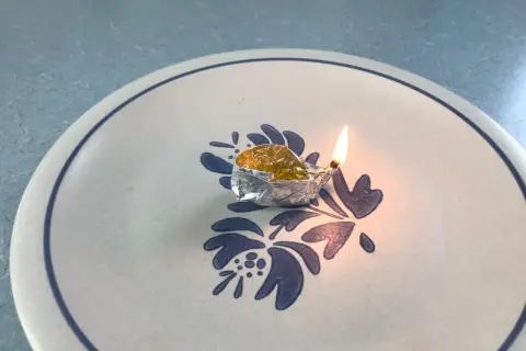 Mała lampka z folii aluminiowej, w której pali się knot zanurzony w naftcie, stoi na talerzu z niebieskim wzorem.