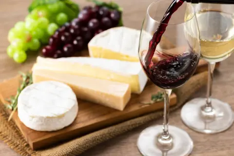 Un bicchiere di vino rosso viene versato, accanto a formaggi assortiti e grappoli d'uva. L'abbinamento perfetto di vino per formaggi.