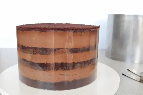 Czekoladowa torta z warstwami ciasta i kremu, przygotowywana w transparentnej formie. Obok stoi metalowy pierścień.