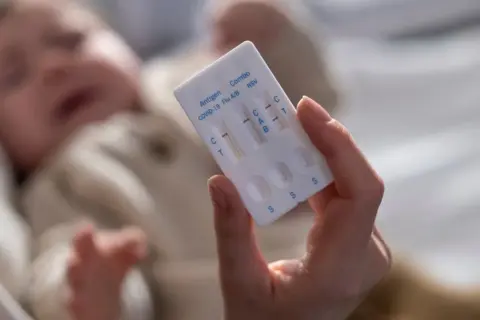 Dłoń trzyma test combo, który wykrywa wirus RSV, grypę i COVID-19. W tle niewyraźne dziecko.