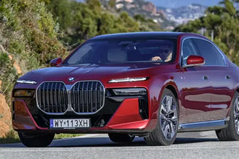 Czerwone BMW 740d z eleganckim czarnym dachem sunie po krętej drodze wśród zielonych wzgórz.