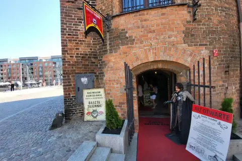 Gdańska Baszta Tajemnic zaprasza do swojego mrocznego świata. Figurka postaci w stroju z epoki stoi przed wejściem.