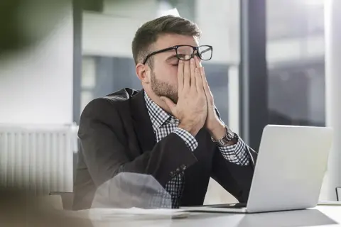 Man med glasögon sitter vid en laptop, gnuggar ögonen. Ett klassiskt dilemma exempel på stressad arbetsdag.