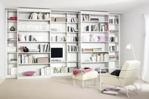 Eine moderne, weiße **eigene Bibliothek** mit vielen Büchern, Dekorationen und einem Fernseher. Ein Sessel und ein Hocker laden zum Verweilen ein.