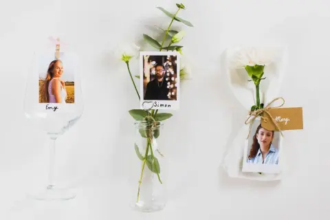 Marque place fleur : photos de invités (Emy, Simon, Mary) attachées à des fleurs et des verres, pour une décoration de table personnalisée.