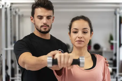 Trener pomaga kobiecie ćwiczyć z hantlem. To przykład, jak fitness co to jest – aktywność fizyczna dla zdrowia i lepszego samopoczucia.