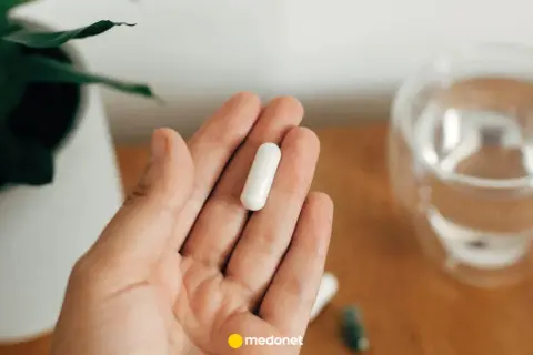 Dłoń trzyma białą kapsułkę. Obok szklanka wody i roślina. Uważaj na nadmiar magnezu objawy.