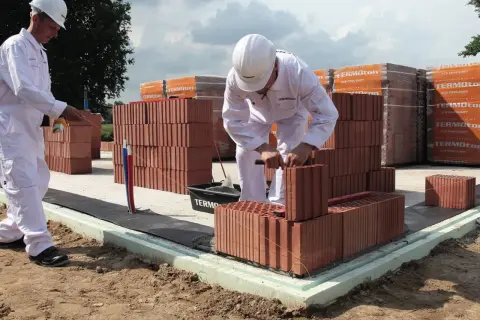Dwóch budowlańców w białych kombinezonach i kaskach układa cegły na fundamencie.