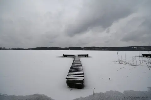 Zimowy pomost na zamarzniętym jeziorze. Ogniskowa 16 mm uchwyciła surowy krajobraz pod pochmurnym niebem.