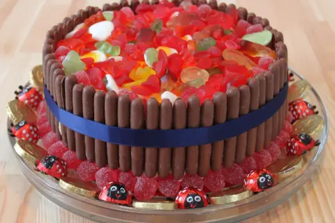 En tårta dekorerad med godis, chokladpinnar och en blå sidenband. Runt tårtan finns godis som ser ut som nyckelpigor och guldfärgade chokladmynt.