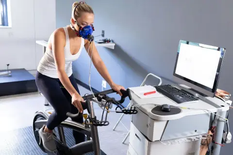 Femme en masque respiratoire effectuant un test d'effort duree sur un vélo d'appartement, connectée à un équipement médical.