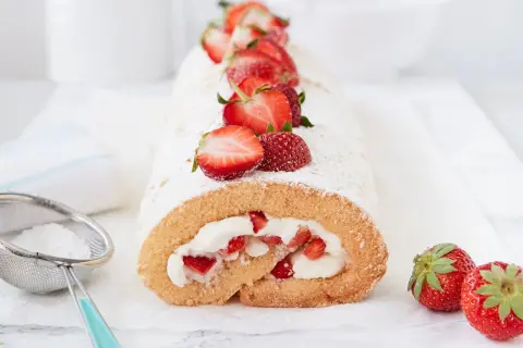 Saftige erdbeer biskuitrolle mit frischen Erdbeeren und Puderzucker bestäubt, daneben ein Sieb mit Puderzucker.