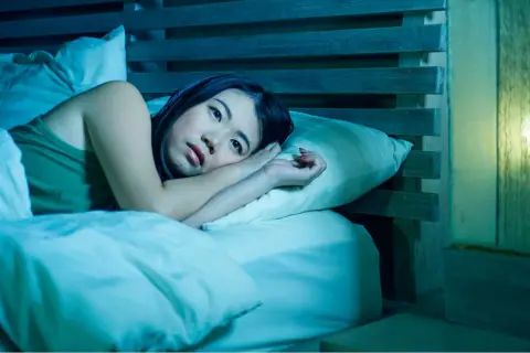 A woman lies awake in bed, her eyes wide open, a picture of paradoxical insomnia. The room is bathed in a cool, blue light, emphasizing her distress.