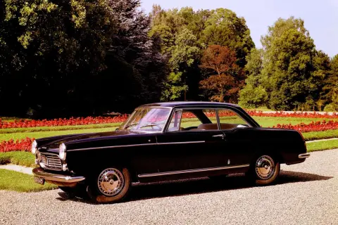 Elegancki, czarny Peugeot 404 coupe z lat 60. stoi na żwirowej alejce, otoczony zielenią i czerwonymi kwiatami.