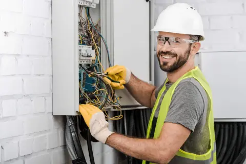 Uśmiechnięty technik elektryk w białym kasku i żółtych rękawicach pracuje przy skrzynce z przewodami.