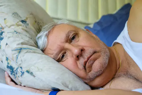 Starszy mężczyzna leży w łóżku, zamyślony. Jego twarz wyraża niepokój, być może związane z cichym udarem i jego objawami.
