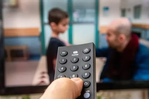 Dłoń trzymająca pilot do telewizora, na ekranie widać scenę z dzieckiem i mężczyzną. Czas na ulubiony serial, opłacony abonament radiowo-telewizyjny działa!