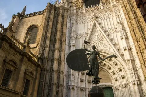 Estatua frente a la imponente fachada de la catedral de Sevilla, con detalles góticos y una escultura que evoca la navegación.