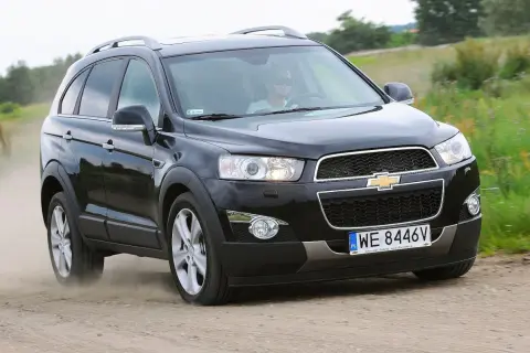 Czarny Chevrolet Captiva jedzie po piaszczystej drodze, wzbijając tumany kurzu. Kierowca w okularach przeciwsłonecznych prowadzi auto przez zielone pola.