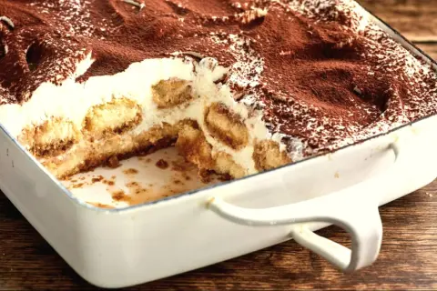 Tiramisu w białej formie, posypane kakao, z widocznymi warstwami biszkoptów i kremu.