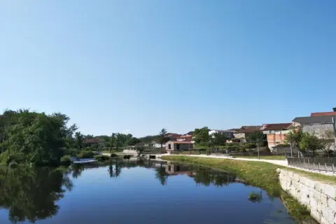 Tranquila playa fluvial en Leiro, con casas tradicionales reflejándose en el agua serena bajo un cielo azul.