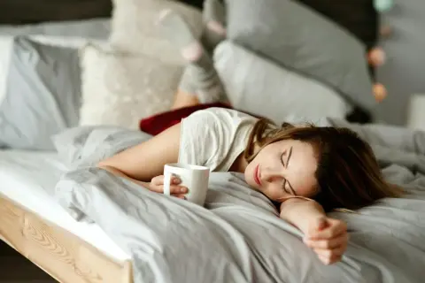 En kvinna sover i sängen med en kaffekopp i handen. Kaffe bra eller dåligt? Kanske en tupplur är bättre.