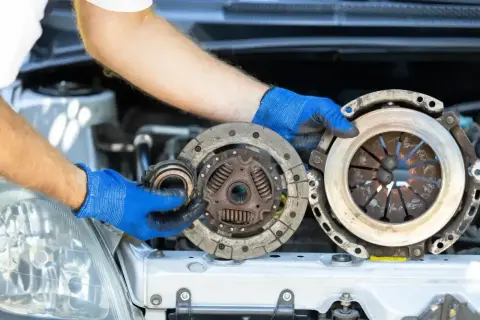 Mechanik w niebieskich rękawiczkach trzyma części samochodowe. Widać zużyte elementy, które mogą powodować ślizgające się sprzęgło.