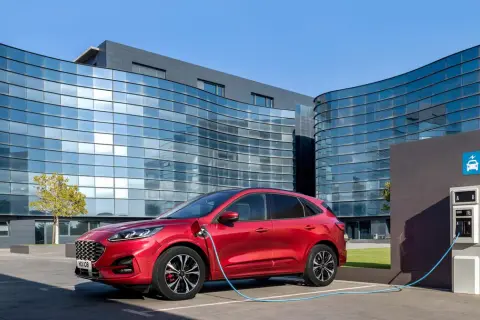 Czerwony Ford Kuga, nowoczesny kombi, ładuje się przy stacji ładowania przed futurystycznym budynkiem.