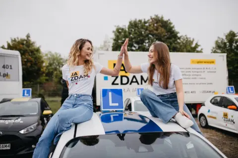 Dwie uśmiechnięte dziewczyny świętują sukces po egzaminie praktycznym kat B. Siedzą na dachu samochodu nauki jazdy, otoczone innymi pojazdami szkoleniowymi.